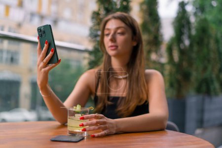 Foto de Una joven sonriente se sienta en una terraza de verano en un bar club con un cóctel y toma una foto con su cámara del teléfono - Imagen libre de derechos