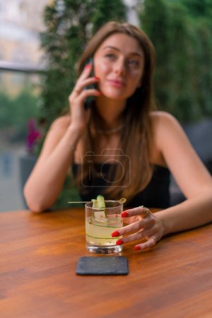 Téléchargez les photos : Jeune fille souriante assise sur une terrasse d'été dans un bar club avec cocktail et parler au téléphone - en image libre de droit