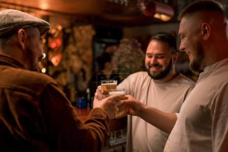 Téléchargez les photos : Gros plan d'un groupe de trois amis debout au bar discutant et trinquant avec des verres de bière et un cocktail au bar du club - en image libre de droit