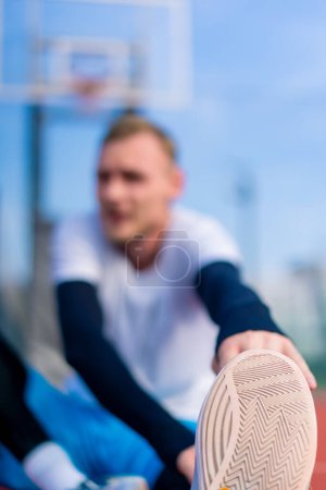 Photo for Tall guy basketball player sits on a basketball court outside and stretches his muscles before the start of practice close-up - Royalty Free Image