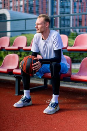 Photo for Tall guy basketball player sits in the bleachers of an outdoor basketball court with ball in hand before practice starts - Royalty Free Image