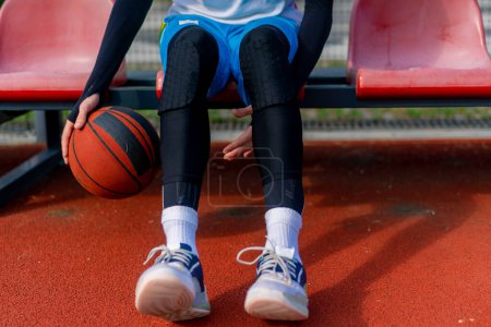 Photo for A tall guy basketball player sits in the bleachers of an outdoor basketball court and demonstrates dribbling basketball before practice starts - Royalty Free Image
