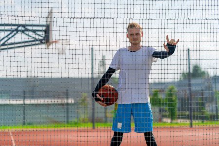 Photo for Tall guy basketball player standing on a basketball court on the street with ball in hand view through the fence netting - Royalty Free Image
