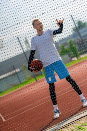 Photo for Tall guy basketball player standing on a basketball court on the street with ball in hand view through the fence netting - Royalty Free Image