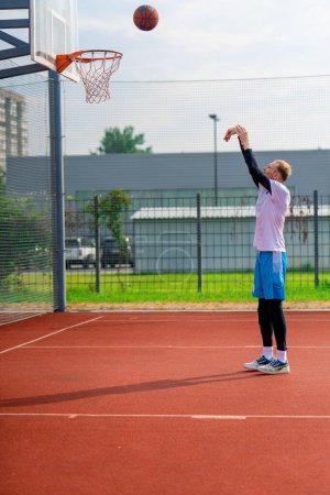 Téléchargez les photos : Grand joueur de basket-ball jette une balle dans un panier de basket-ball sur un terrain de basket-ball dans le parc pendant l'entraînement - en image libre de droit