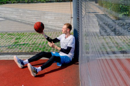Photo for A tall guy basketball player with a ball in his hand sits leaning on the net of a basketball court during practice in park - Royalty Free Image