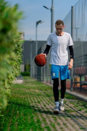 Photo for Tall guy basketball player walks down the park path to the basketball court and drives the ball showing off his dribbling skills - Royalty Free Image