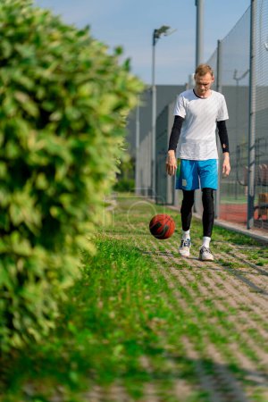 Photo for Tall guy basketball player walks down the park path to the basketball court and drives the ball showing off his dribbling skills - Royalty Free Image