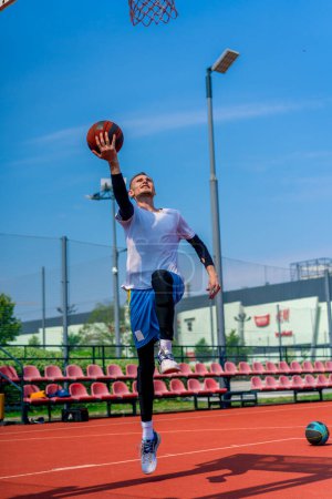 Foto de Alto jugador de baloncesto salta al aro con la pelota en la mano para anotar una espectacular inmersión durante la práctica en la cancha de baloncesto en el parque - Imagen libre de derechos