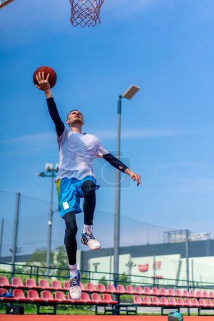 Photo for Tall guy basketball player jumps to the hoop with the ball in his hand to score a spectacular dunk during practice on the basketball court in park - Royalty Free Image