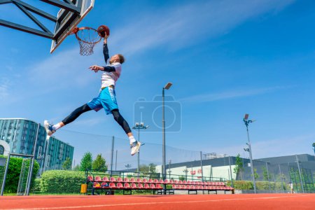 Foto de Alto jugador de baloncesto salta al aro con la pelota en la mano para anotar una espectacular inmersión durante la práctica en la cancha de baloncesto en el parque - Imagen libre de derechos