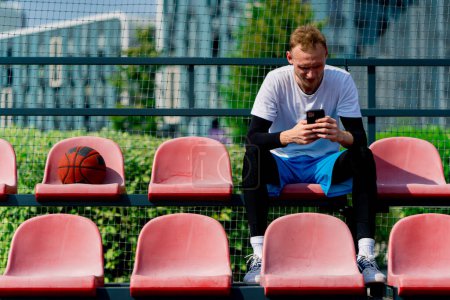 Photo for Tall guy basketball player sits staring into a smartphone on the bleachers of basketball court next to the ball - Royalty Free Image
