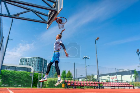 Téléchargez les photos : Grand joueur de basket-ball saute au cerceau avec la balle à la main pour marquer un dunk spectaculaire pendant l'entraînement sur le terrain de basket dans le parc - en image libre de droit