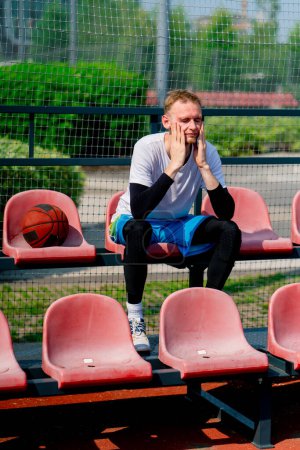 Photo for Tall frustrated guy basketball player sitting in the bleachers of a basketball court with ball by his side - Royalty Free Image