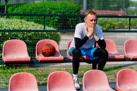 Foto de Alto jugador de baloncesto frustrado tipo sentado en las gradas de una cancha de baloncesto con pelota a su lado - Imagen libre de derechos