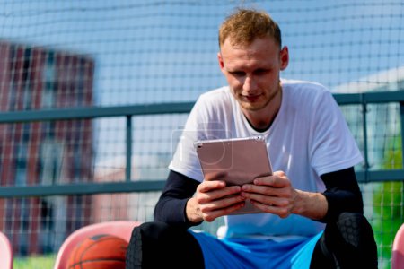 Téléchargez les photos : Un grand joueur de basket-ball s'assoit en regardant une tablette dans les gradins d'un terrain de basket-ball avec une balle à côté de lui - en image libre de droit