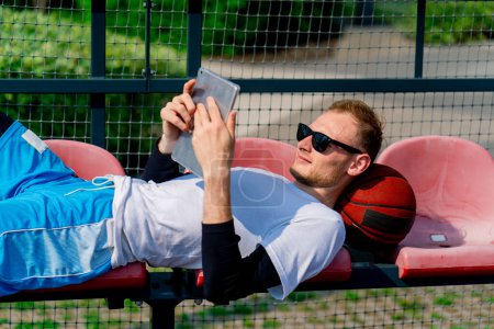 Photo for Tall guy basketball player lays his head on the ball and stares into a clipboard in the bleachers of basketball court - Royalty Free Image