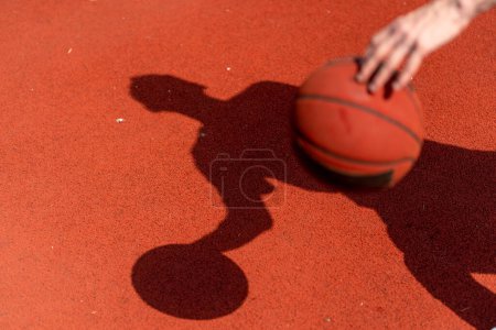 Téléchargez les photos : Gros plan de l'ombre du joueur de basket sur le sol d'un terrain de basket tout en dribblant la balle - en image libre de droit