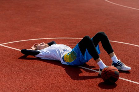 Téléchargez les photos : Grand joueur de basket-ball couché sur un terrain de basket-ball dans le parc avec le basket-ball se reposant pendant l'entraînement - en image libre de droit