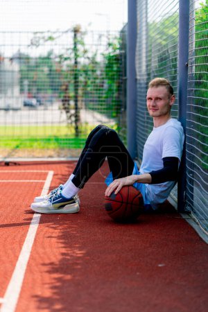 Photo for A tall guy basketball player with a ball in his hand sits leaning on the net of a basketball court during practice in park - Royalty Free Image