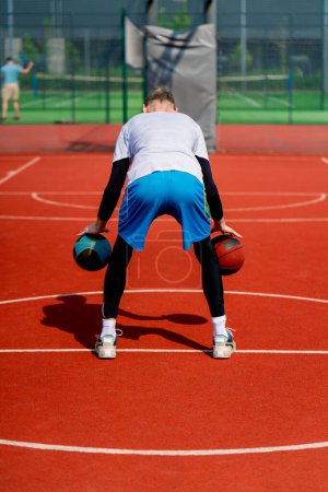 Photo for A tall guy basketball player with two balls shows off his dribbling skills while practicing on basketball court in the park - Royalty Free Image