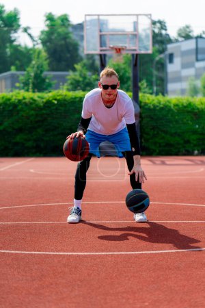 Photo for A tall guy basketball player with two balls shows off his dribbling skills while practicing on basketball court in the park - Royalty Free Image
