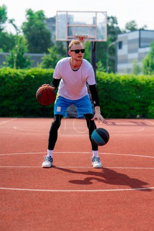Téléchargez les photos : Un grand joueur de basket-ball avec deux balles montre ses compétences de dribble tout en pratiquant sur le terrain de basket dans le parc - en image libre de droit