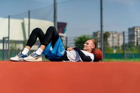 Photo for Tall guy basketball player lying on a basketball court in the park along with  basketball resting during practice - Royalty Free Image