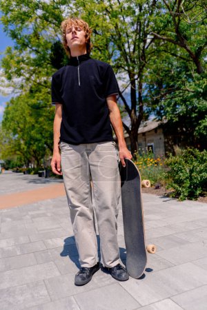 Foto de Retrato de un joven patinador serio con el pelo largo sosteniendo un monopatín en la mano sobre un fondo de skatepark y cielo - Imagen libre de derechos