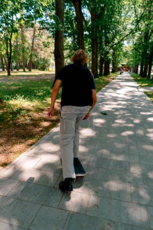 Foto de Joven patinador paseos monopatín en el camino del parque de la ciudad contra el fondo de los árboles y el cielo - Imagen libre de derechos