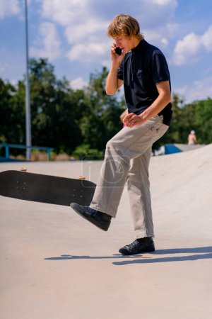 Téléchargez les photos : Un jeune skateboarder aux cheveux longs tenant un skateboard à la main et parlant au téléphone au skatepark de la ville - en image libre de droit