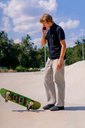 Foto de Un joven patinador con el pelo largo sosteniendo un monopatín en la mano y hablando por teléfono en el parque de skate de la ciudad - Imagen libre de derechos