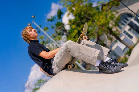 Photo for A young guy skateboarder with long hair sits on his skateboard on the edge of a skate pool at city skatepark - Royalty Free Image