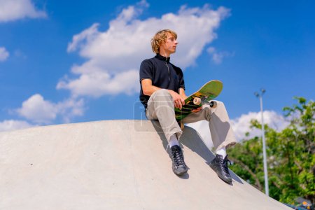 Foto de Un joven patinador con el pelo largo se sienta en su monopatín en el borde de una piscina de skate en el parque de skate de la ciudad - Imagen libre de derechos