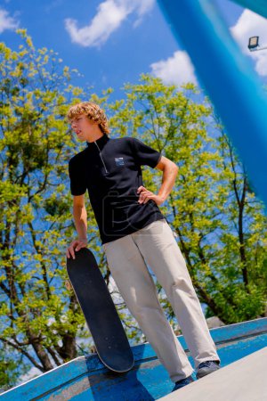 Téléchargez les photos : Un jeune skateboarder avec les cheveux longs et une planche à roulettes dans sa main se tient près des mains courantes tout en se reposant entre les tours au skatepark de la ville - en image libre de droit