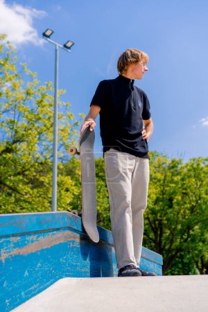 Foto de Un joven patinador con el pelo largo y un monopatín en la mano se para cerca de pasamanos mientras descansa entre trucos en el parque de skate de la ciudad - Imagen libre de derechos