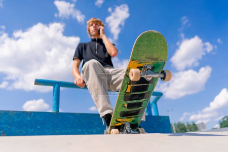 Téléchargez les photos : Un jeune skateboarder assis sur un skateboard avec des mains courantes et parlant au téléphone dans un skatepark de la ville - en image libre de droit