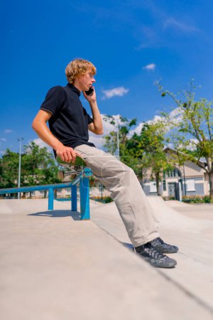 Foto de Un joven patinador sentado en un monopatín con barandillas y hablando por teléfono en el parque de skate de la ciudad - Imagen libre de derechos