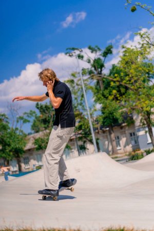Foto de Joven skateboarder con pelo largo skateboarding en el parque de skate de la ciudad y hablando por teléfono - Imagen libre de derechos