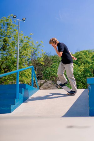 Foto de Joven skateboarder con pelo largo skateboarding en el parque de skate de la ciudad y hablando por teléfono - Imagen libre de derechos