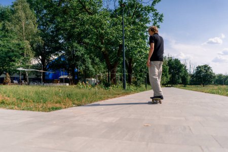 Photo for Young guy skater rides skateboard on the path of the city park against the background of trees and sky - Royalty Free Image