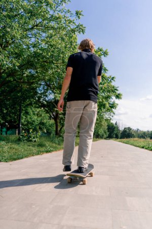Foto de Joven patinador paseos monopatín en el camino del parque de la ciudad contra el fondo de los árboles y el cielo - Imagen libre de derechos