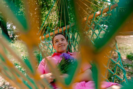 Téléchargez les photos : Jeune fille couchée sur hamac dans le parc reposant de roller et tenant un bouquet de fleurs dans ses mains modèle plus-taille - en image libre de droit