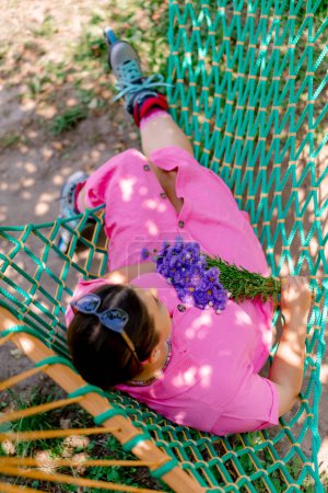 Foto de Una chica en un vestido rosa con patines de ruedas con un ramo de flores en las manos está acostada en una hamaca en el parque - Imagen libre de derechos