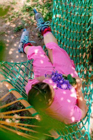 Téléchargez les photos : Une fille dans une robe rose portant des patins à roulettes avec un bouquet de fleurs dans ses mains est couchée sur un hamac dans le parc - en image libre de droit