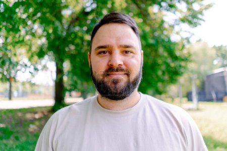 Téléchargez les photos : Portrait d'un homme souriant avec une barbe dans un t-shirt blanc debout dans un parc sur un fond d'arbres plus un modèle de taille - en image libre de droit