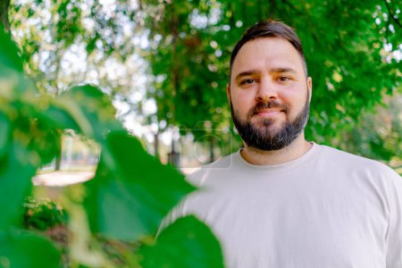 Téléchargez les photos : Portrait d'un homme souriant avec une barbe dans un t-shirt blanc debout dans un parc sur un fond d'arbres plus un modèle de taille - en image libre de droit