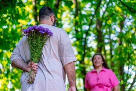 Foto de Joven pareja hombre se encuentra con chica en el parque y esconde un ramo de flores detrás de su espalda más el modelo de tamaño - Imagen libre de derechos