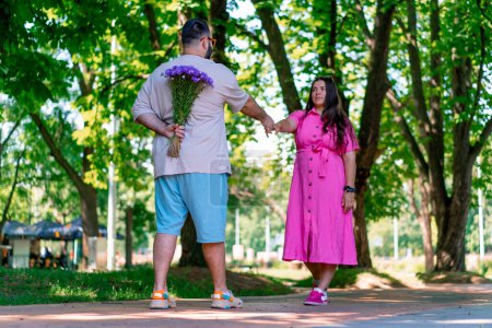 Photo for Young couple man meets girl in the park and hides a bouquet of flowers behind his back plus size model - Royalty Free Image