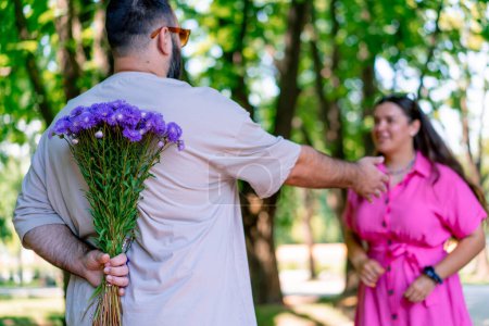 Foto de Joven pareja hombre se encuentra con chica en el parque y esconde un ramo de flores detrás de su espalda más el modelo de tamaño - Imagen libre de derechos